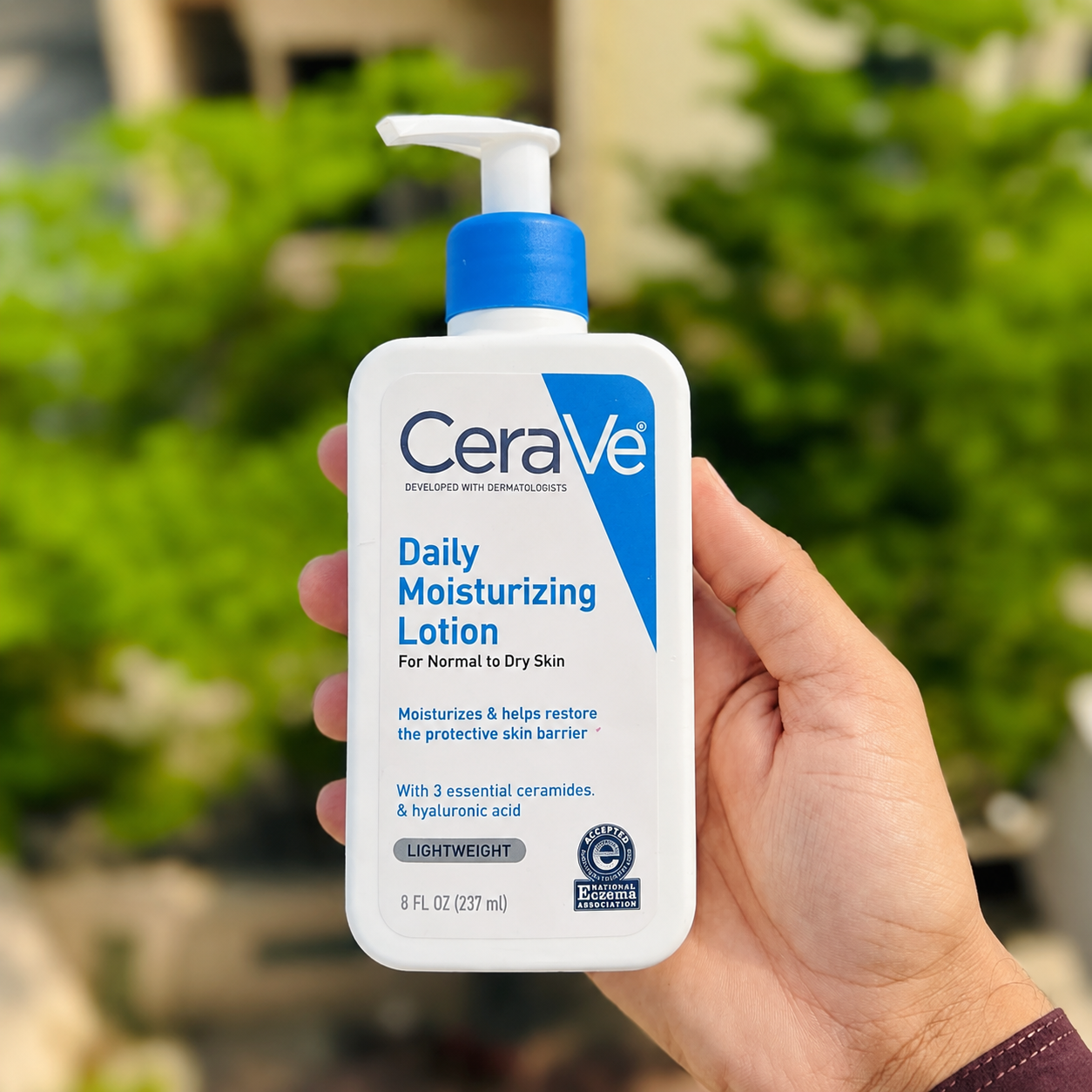 A hand holding a bottle of CeraVe Daily Moisturizing Lotion against a blurred green outdoor background.