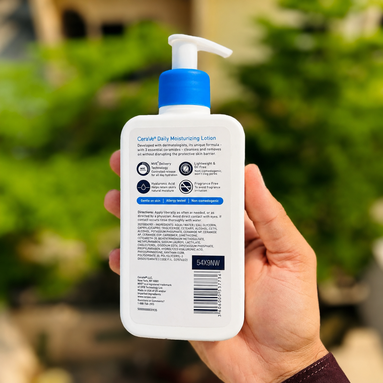 A hand holding a white bottle of Carvyl Daily Moisturising Lotion, showing the back of the packaging with blue pump and blue text, against a blurred green garden background.